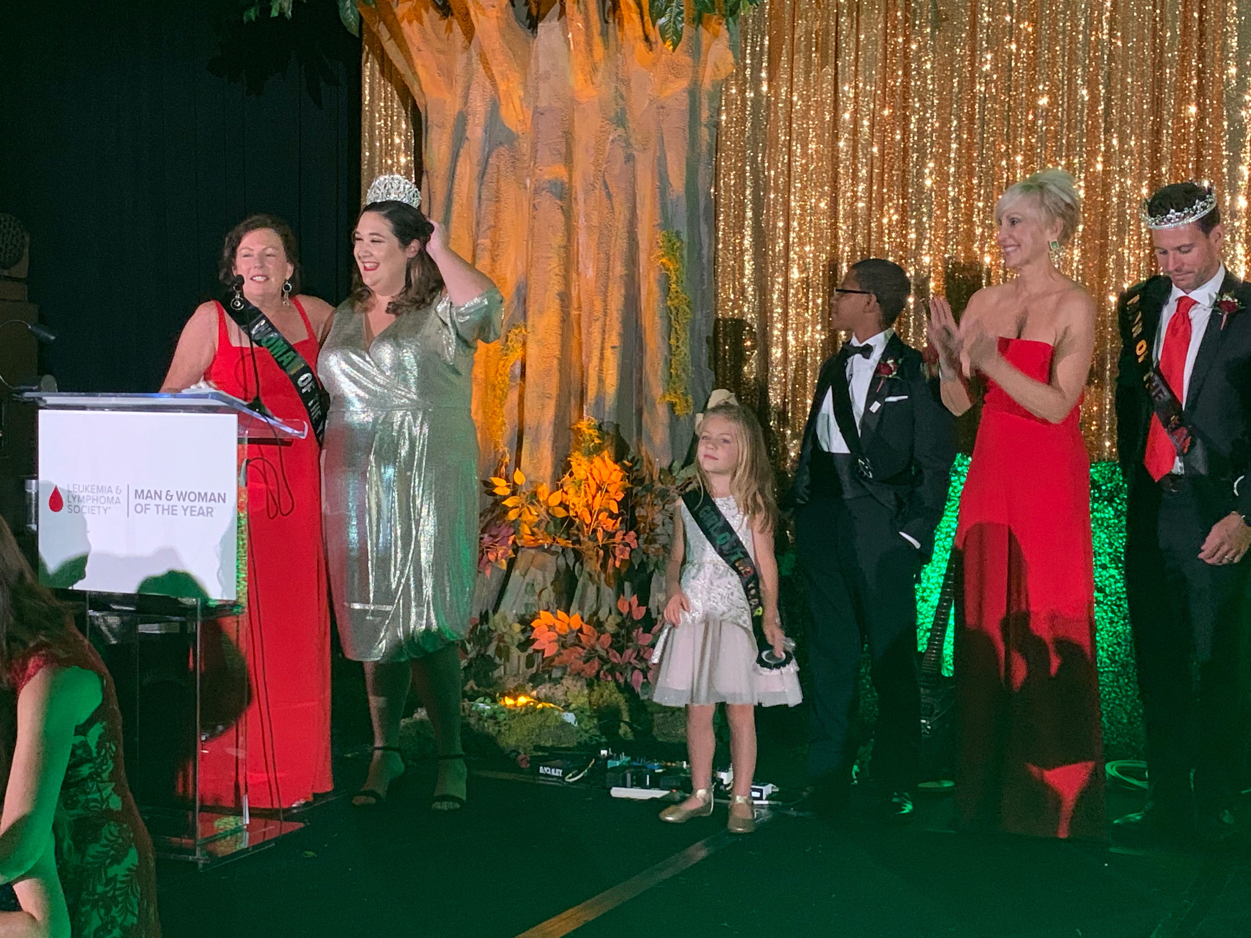Camille Olivere (at podium in red) accepting the honor and crowning her campaign manager, Sara Blakeslee. Standing by are the 2019 Boy & Girl of the Year, 2018 Woman of the Year winner Michelle Homoky of Celebrity Cruises, and 2019 Man of the Year winner Clayton Idle. 